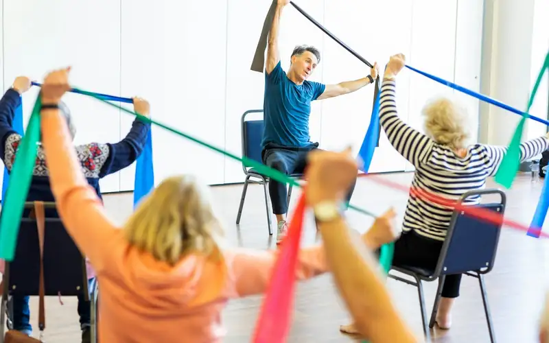 Groep ouderen doet zittend oefeningen met elastieken banden, met een begeleider voorin